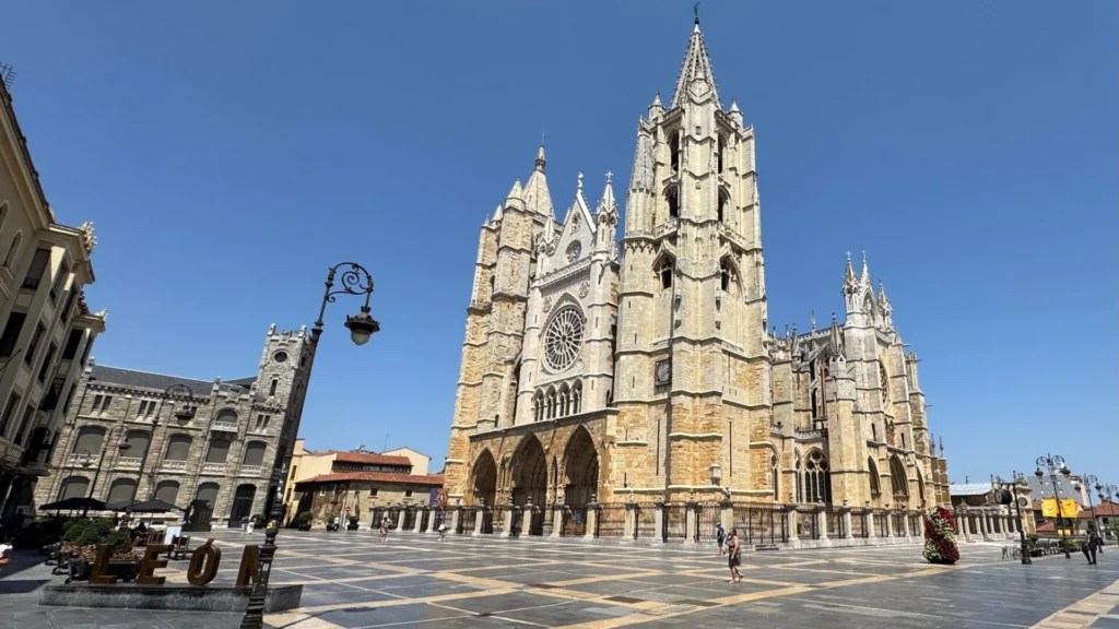 The Gothic Cathedral of León in Spain on a bright sunny day, featuring twin spires, an ornate rose window, pointed arches, and intricate stone tracery, standing majestically in the main square surrounded by historic buildings and visitors