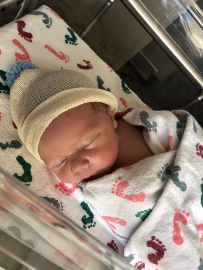 A baby boy in the hospital metal and clear plastic crib wearing a yellow hat with a blue Pom Pom. He is swaddled in a white and footprint patterned hospital blanket.