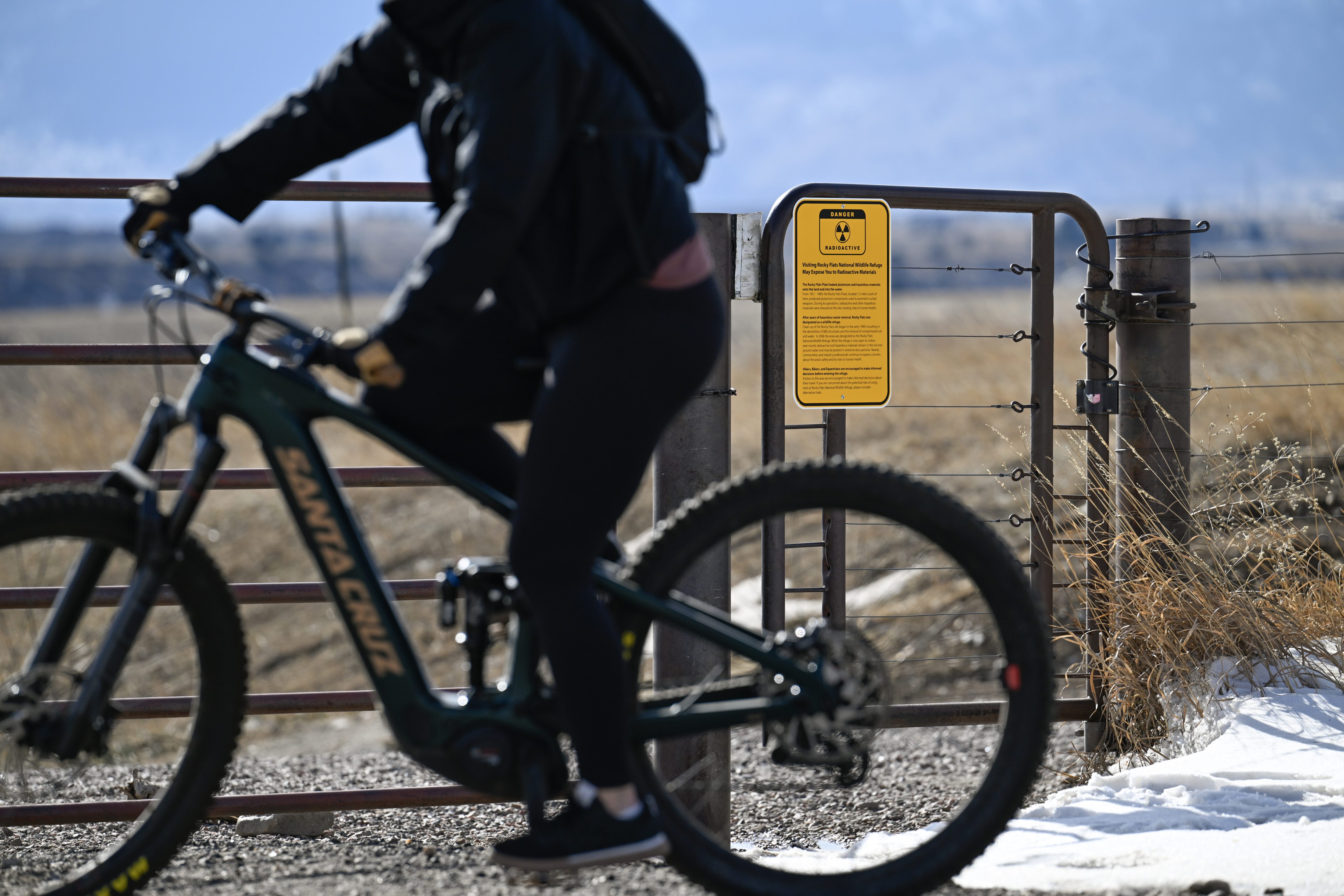 A cyclist rides past a warning sign about radioactive materials on a gate leading to the Colorado 128 underpass at the Rocky Flats National Wildlife Refuge in January 2026. The sign was posted in December "in response to requests from members of the public for additional signage," a county spokesperson said. (Matthew Jonas/Staff Photographer)