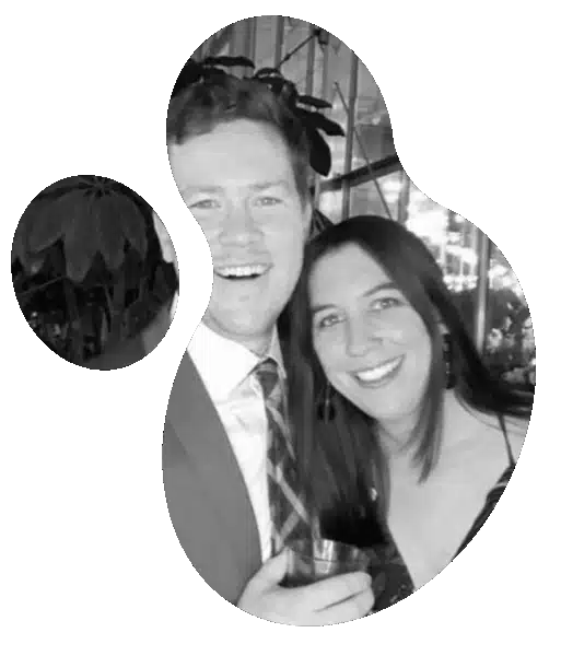 Two people posing indoors at a social event, one in a suit with a checkered tie holding a drink, the other in a dark top, with plants and a window in the background.