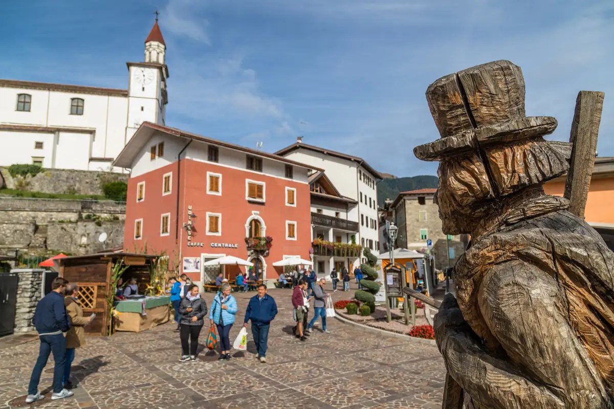 ForMandi a Sutrio con visitatori nella piazza del borgo, la chiesa e una scultura in legno in primo piano