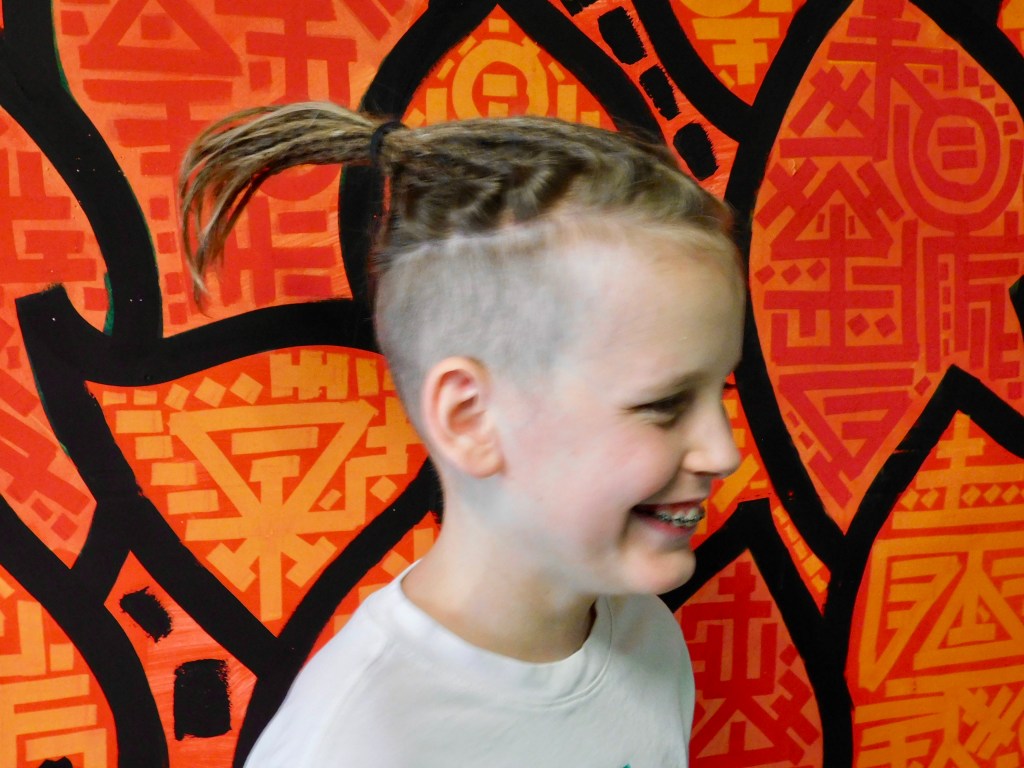 This picture shows a 12-year-old child with partial crochet dreadlocks on top of his head. He is smiling. The background is a red, orange and black mural.