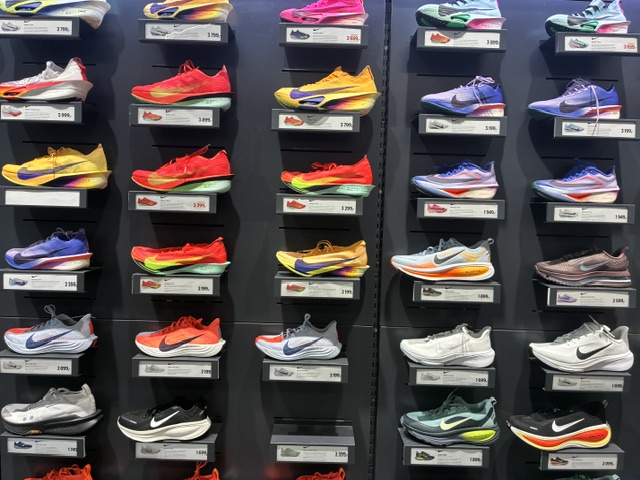 Utstilling av moderne Nike løpesko på vegg i sportsbutikk. Mange av skoene har tykke mellomsåler, rocker-form og fargerike design, typisk for dagens premium løpesko.