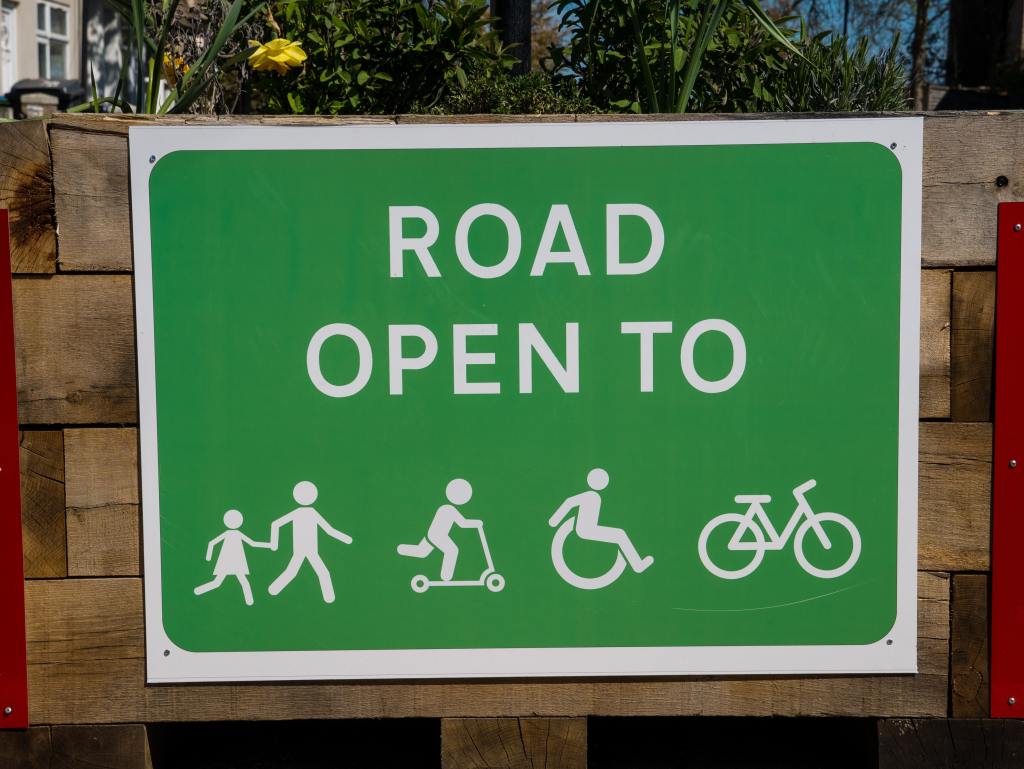 A freen road sign on a planter reading 'Road open to' followed with icons representing children playing, bikes and wheelchair users