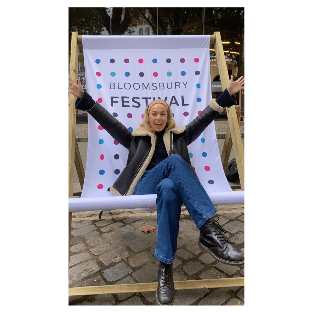 A smiling woman, with her arms outstretched, sitting on a large deckchair emblazoned with the words 'Bloomsbury Festival'
