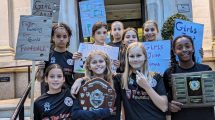 A group of young girls in football kits with placards and a trophy, standing on the steps of Islington Town Hall
