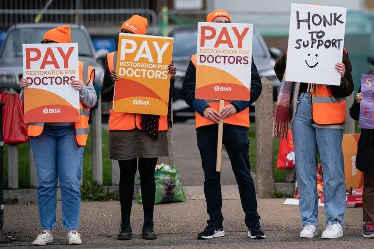 4 people standing in a row with placards that say 'Pay Restoration for Doctors'