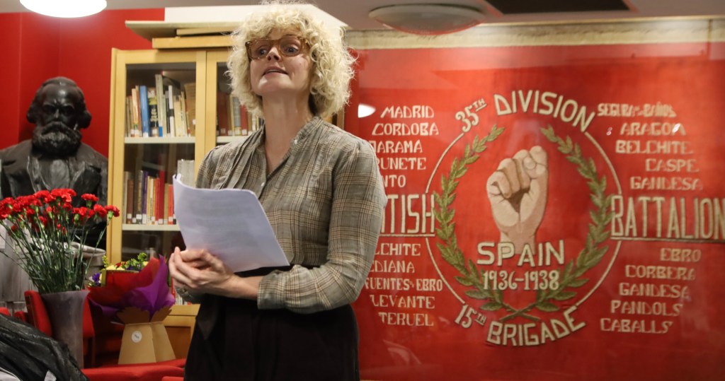 A woman standing in front of a red workers brigade banner, reading from a piece of paper