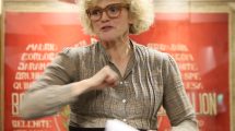 A woman with blonde curly hair and glasses speaking in front of a red banner