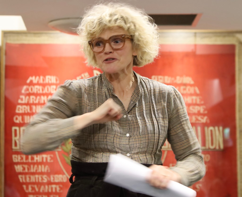 A woman with blonde curly hair and glasses speaking in front of a red banner