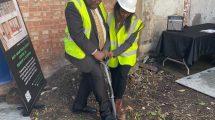 Two people with construcion helmets and hi-vis jackets on put a foot each on a spade as if about to dig the ground