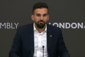 A man with a beard, looking serious, in front of a wall with London Assembly written on it