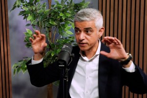 A speaker gestures animatedly while seated in front of a microphone, with a decorative plant and wooden backdrop visible.
