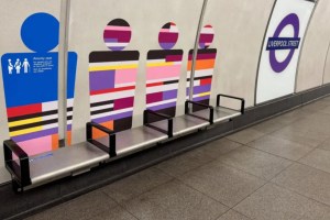 A bench near colourful wall art depicting stylised figures, labelled “Priority seat” for passengers needing assistance, at Liverpool Street station.
