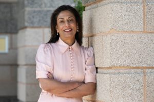 A person in a light pink blouse with puff sleeves stands with arms crossed against a stone wall, exuding a calm and confident demeanour.