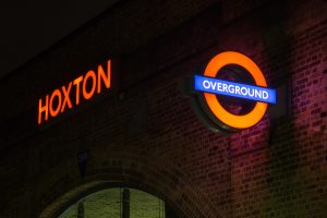 A nightime view of the Hoxton train station sign, featuring bright red "HOXTON" lettering and a blue "OVERGROUND" sign with an orange circle.