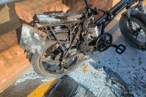 A damaged electric bike with burnt wires, blackened parts, and a charred battery, resting on a brick path.