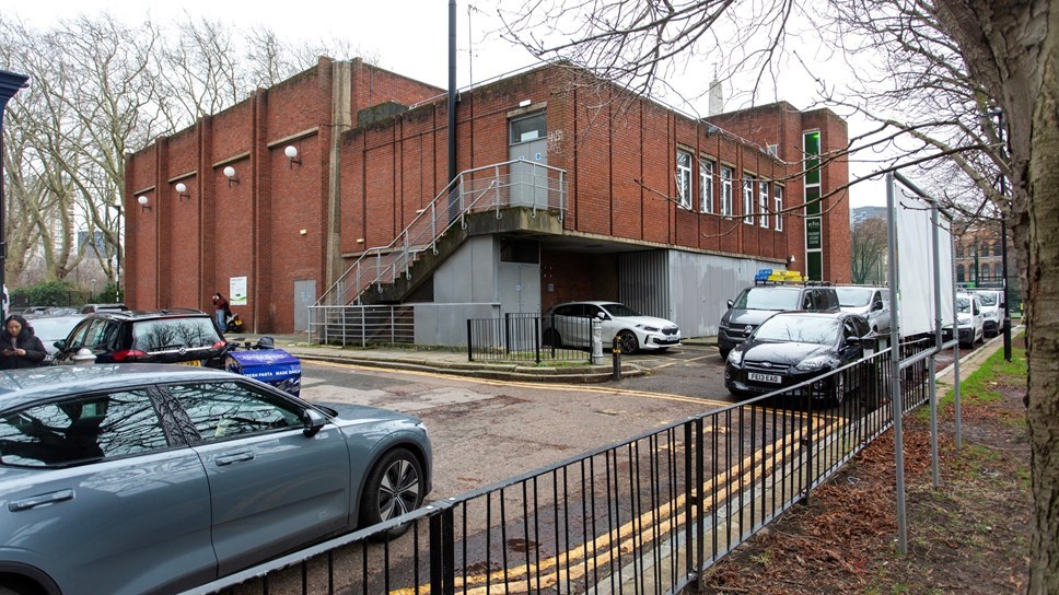 A brick building with a staircase, surrounded by parked cars on a city street, trees in the background, on a cloudy day.