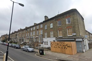 Street view of a row of Victorian-style houses, some boarded up, with graffiti on the wooden barrier, under a cloudy sky.