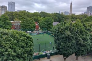 A view of a green football pitch surrounded by trees, with players engaged in a game and city buildings in the background under a cloudy sky.