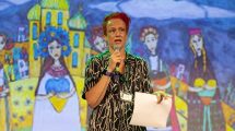 A speaker stands on stage holding a paper and a microphone, with a colorful mural depicting women and buildings in the background.