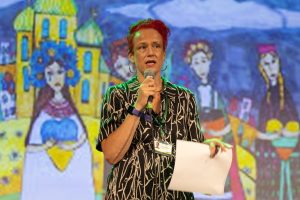 A speaker stands on stage holding a paper and a microphone, with a colorful mural depicting women and buildings in the background.