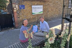 Two people sit on a wooden bench outside Robert Blair Primary School, holding a certificate, with flowers in the foreground.