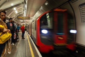 A busy London Underground station with commuters waiting as a train approaches, illuminated signs displaying station information.