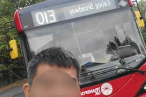 A red bus with "E10 Ealing Broadway" displayed on the front, parked near trees on a cloudy day. A person gestures in front of it.