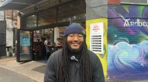 A person with long dreadlocks stands in front of a cafe, with colourful mural art and a sign for the Dusty Knuckle bakery visible.