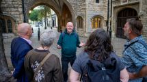 A group of four people attentively listen to a speaker near an ancient stone archway in a quaint street setting.