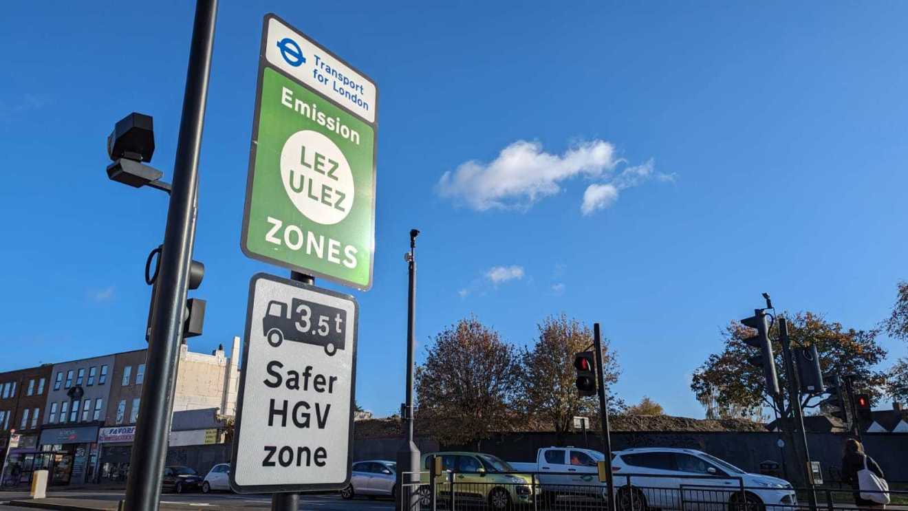Traffic signs indicating the Low Emission Zone (LEZ) and Safer HGV Zone in London under a clear blue sky.