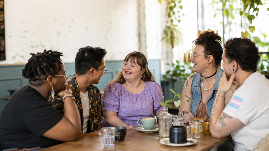 Diverse group of LGBTQ+ adults, including a woman with Down syndrome, sharing conversation and laughter at a café.