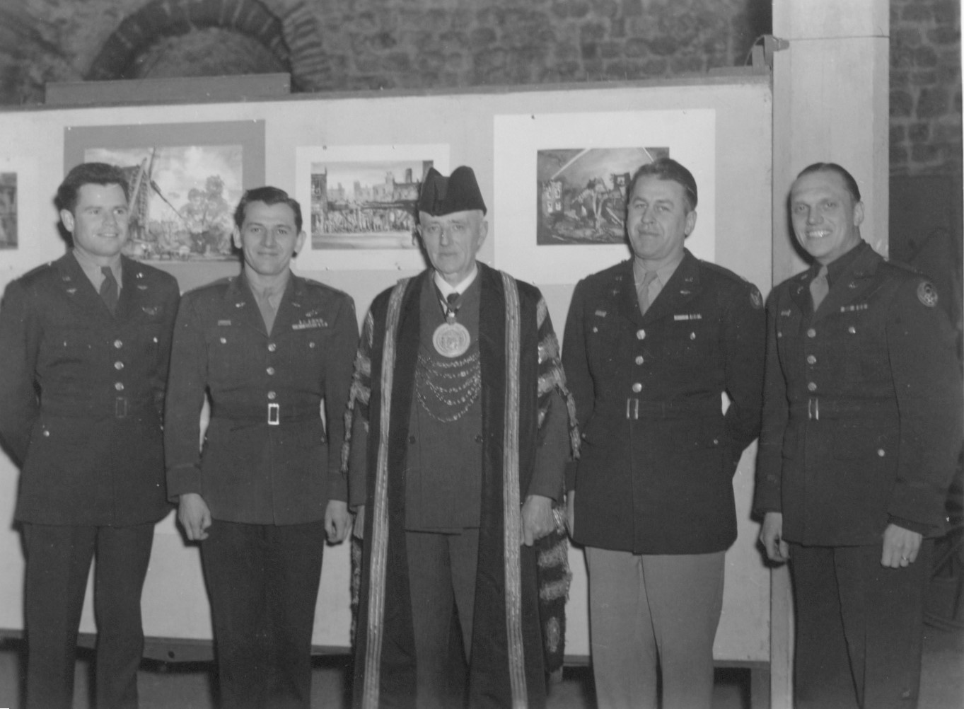 Four men in uniform stand around a man in ceremonial robes. Behind them is an exhibition of artwork and a stone wall behind that.