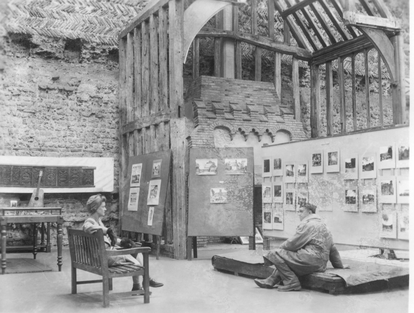Black and white photograph of a stone building with wooden timber structure and exhibition panels. A woman sits on a bench and a man sits on a pallet in the foreground.