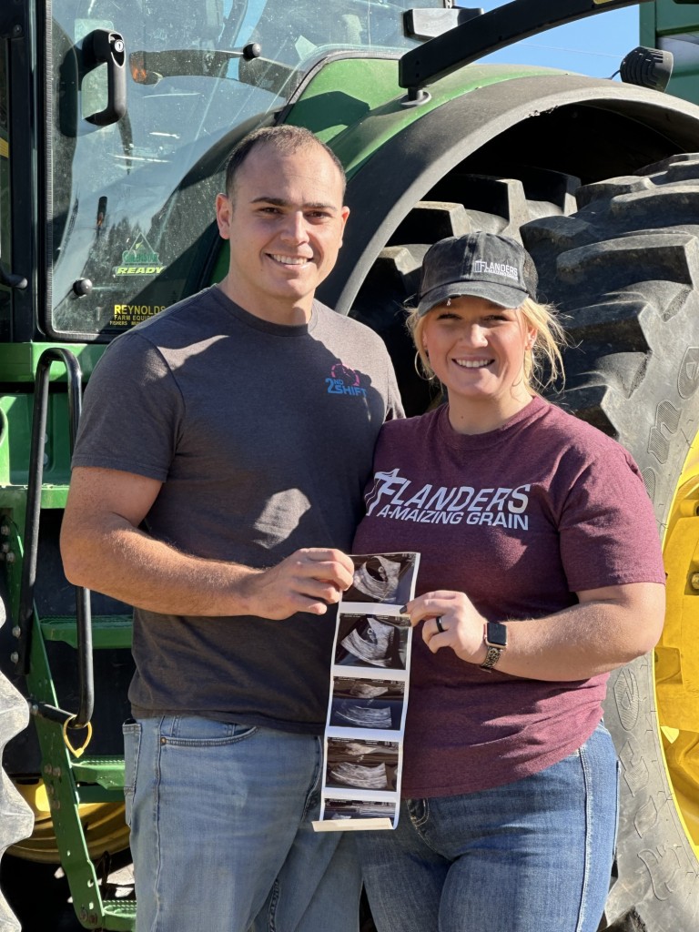 Expecting farm couple holding ultrasound pictures in front of John Deere tractor