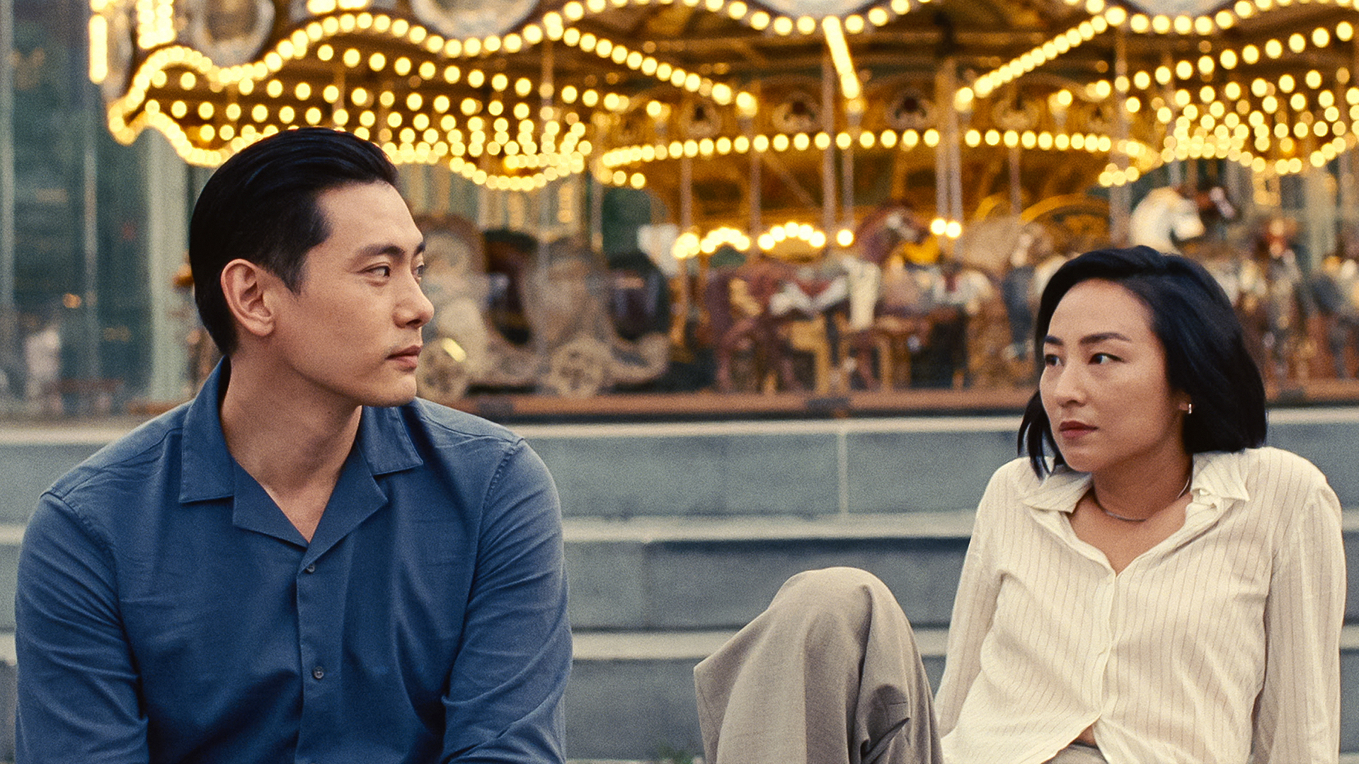 Hae Sung (Teo Yoo) and Nora (Greta Lee) sitting in front of a carousel.