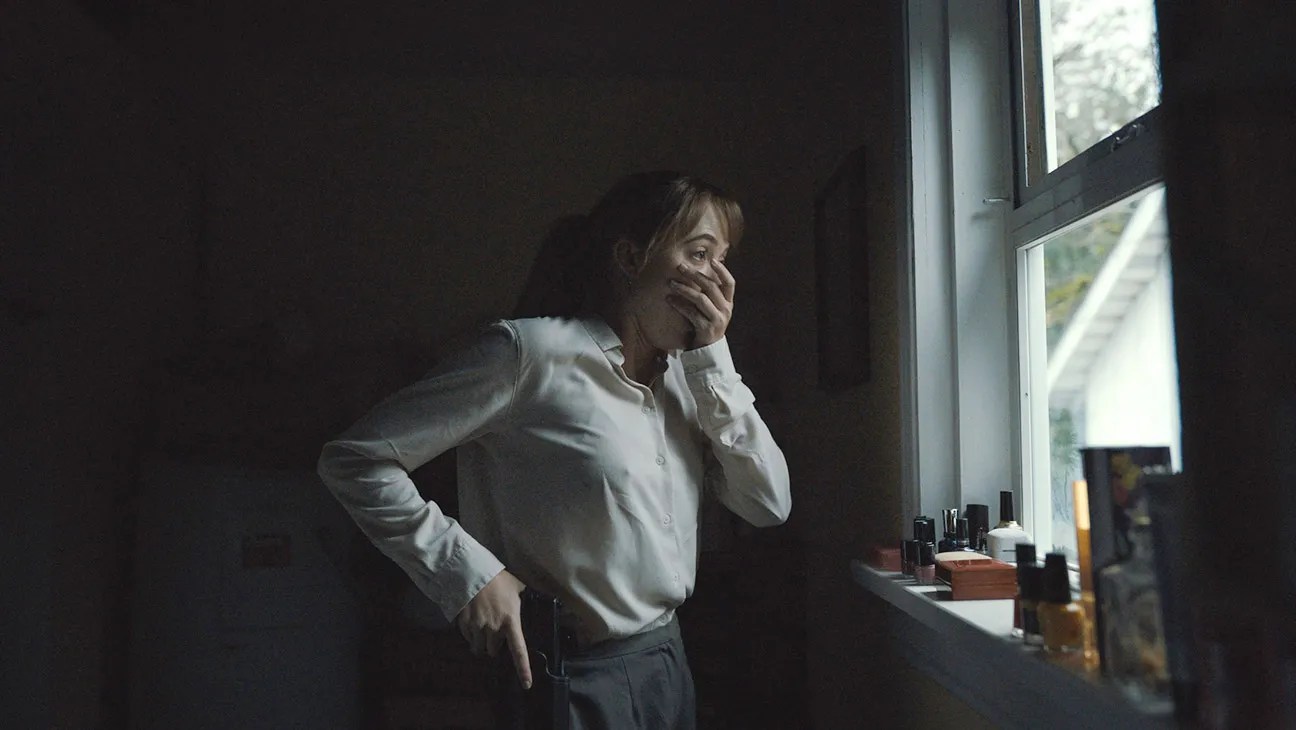 Lee stands in front of a window with one hand covering her mouth in fear and the other on her gun.
