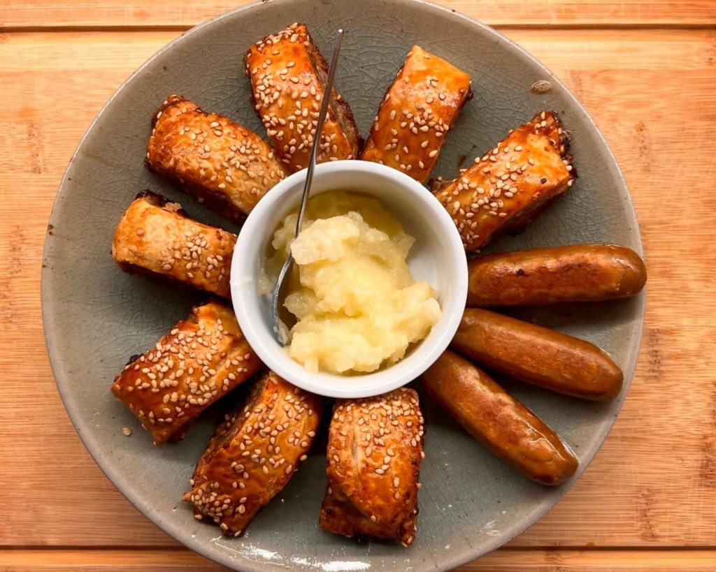 plate of gluten-free sausage rolls and vegan sausages, arranged in a ring around a small pot of apple sauce.