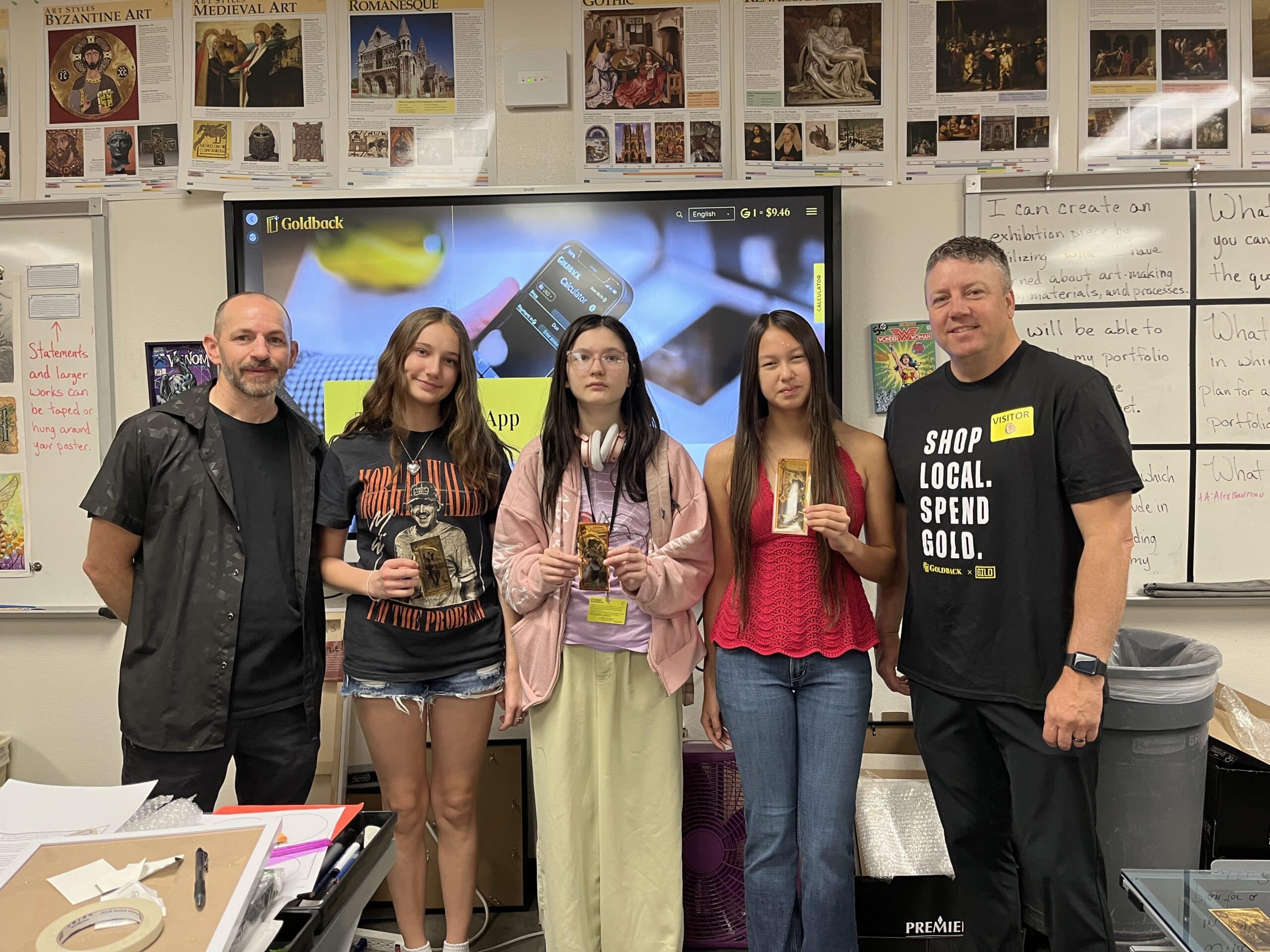 A group of five people stands in an art classroom in front of a screen displaying the Goldback website. Three students in the center hold Goldback notes, with two adults standing on either side. Art history posters and whiteboards fill the wall behind them