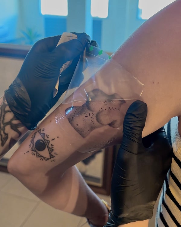 tattoo artist removes the top layer of the second skin from a fresh tattoo. this is the first part of the healing process. The tattoo seen under the clear adhesive is of Aphrodite, classical statue in dotwork style.