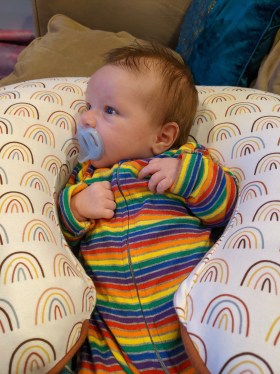 Baby Metra sits in the middle of a round cushion watching TV while wearing rainbow pajamas and sucking on a pacifier.