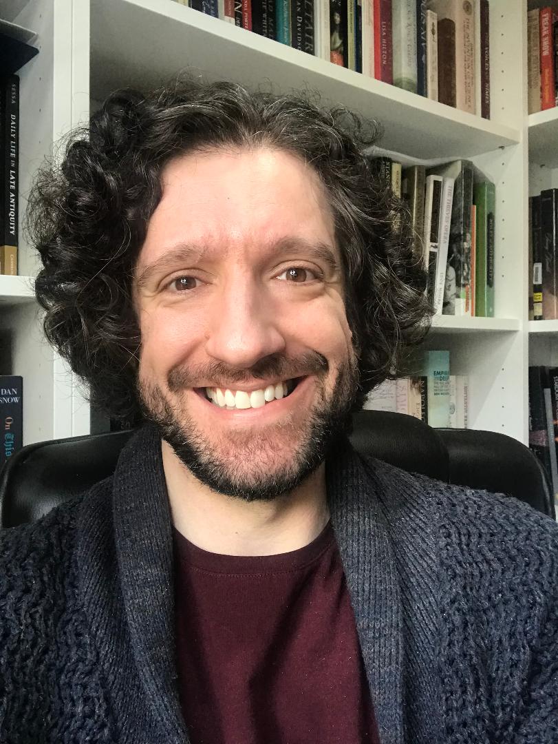 Photo of Greg Jenner smiling. He has curly brown hair, a short beard, and wears a maroon t-shirt under a blue knitted cardigan