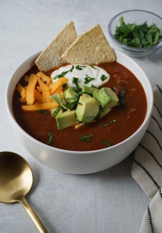 Stovetop Chili in a white bowl with avocado and cheese and sour cream on top with a cold spoon