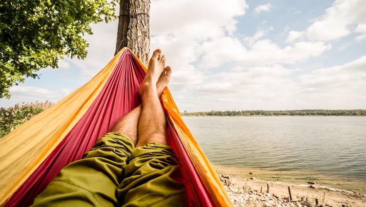 Relaxing in the hammock at the beach under a tree, summer day. Barefoot man laying in hammock, looking on a lake, inspiring landscape