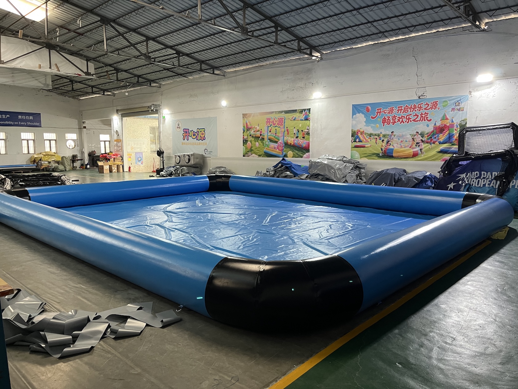 Interior view of blue inflatable water pool showing the PVC floor base and black corner supports in a warehouse setting