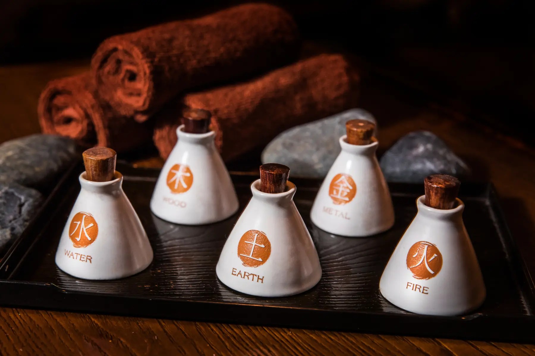 Five small ceramic aromatherapy bottles labeled Water, Wood, Earth, Metal, and Fire sit on a black tray, with rolled towels and smooth stones in the background.