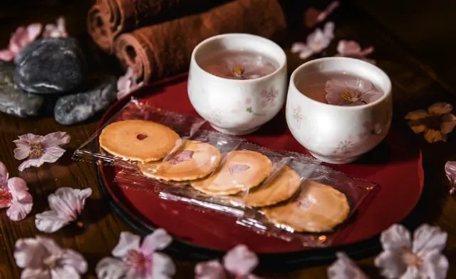 Two cups of tea with cherry blossom flowers and five packaged cherry blossom cookies are arranged on a red tray, surrounded by pink flower petals and decorative stones.