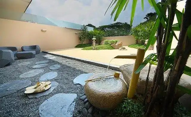 A minimalist Japanese-style garden with a stone water fountain, bamboo, stepping stones, sandals, greenery, and outdoor seating under an open sky.