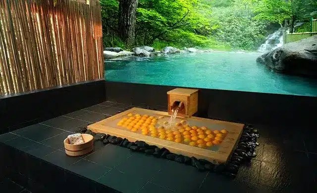 A wooden indoor bath filled with floating oranges sits next to a large open-air hot spring surrounded by lush green trees and rocks.